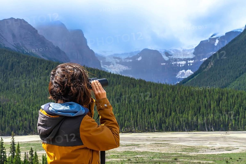 Tourist wearing an orange jacket uses binoculars to observe a glacier in the canadian rockies, surrounded by a coniferous forest and mountains