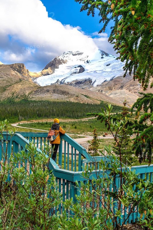 Tourist with backpack standing on wooden turquoise viewpoint is admiring stunning view of athabasca glacier in jasper national park, alberta, canada