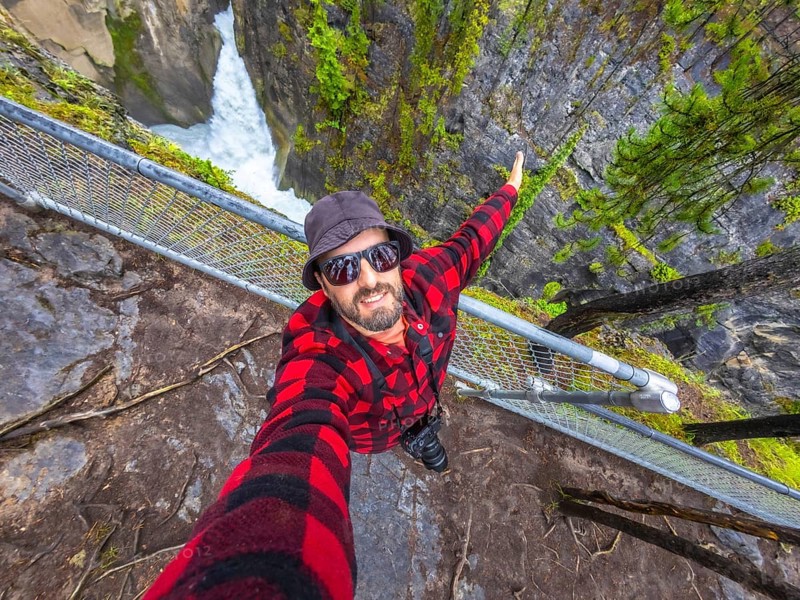 Photographer wearing sunglasses and a bucket hat takes a selfie at sunwapta falls in jasper national park, extending one arm for a wider view of the waterfall and surrounding forest