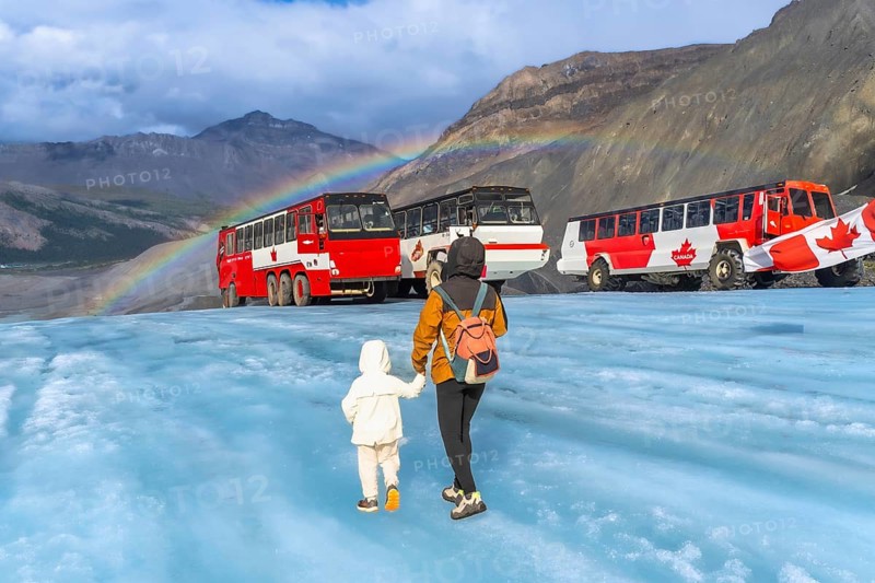 Tourists walking on the surface of athabasca glacier in jasper national park, alberta, canada, with large red tour buses parked nearby and a vibrant rainbow arching across the sky