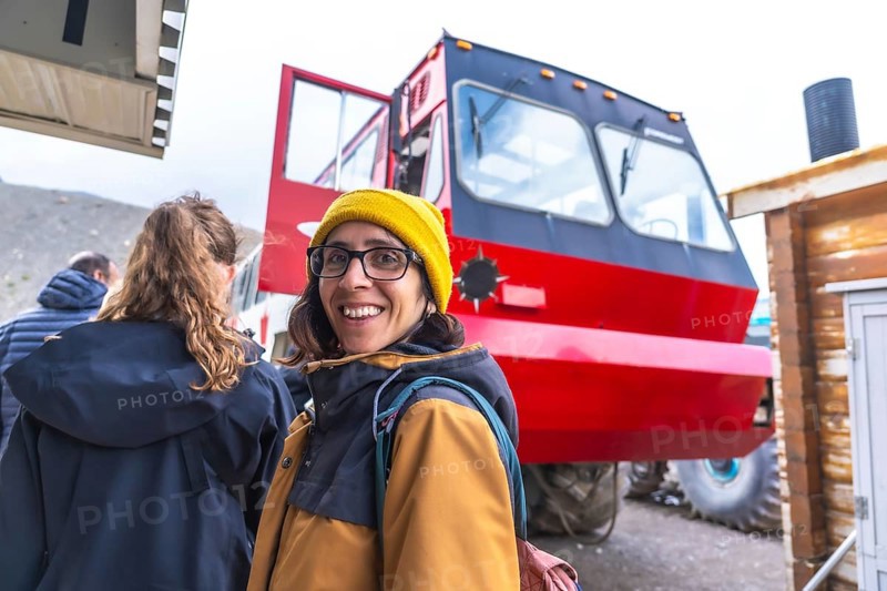 Tourist wearing a yellow beanie and glasses smiles near a large red bus, used to transport visitors onto the athabasca glacier in jasper national park, alberta, canada