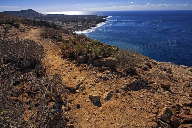 A rocky path leads along the coast, the sea shines in the distance under a clear sky, Los Cristianos Tenerife