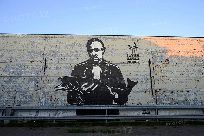 A large mural shows Marlon Brando in the Godfather holding a fish. Mural by street artist Pobel. The inscription means salmon is important for Norway. The art is in black and white and looks rough and urban, Vardø, Finnmark, Norway
