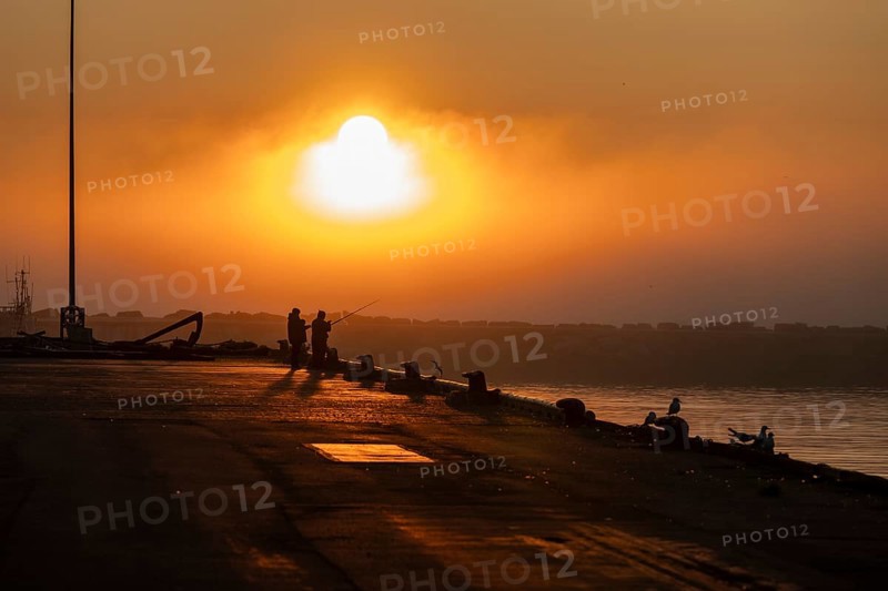 Silhouettes of people fishing at harbour at sunset under a warm orange sky, Vardø, Finnmark, Norway