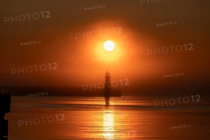A lighthouse at the Vardö pier stands silhouetted against an orange sunset over the sea reflecting the colors of the sky, Vardø, Finnmark, Norway