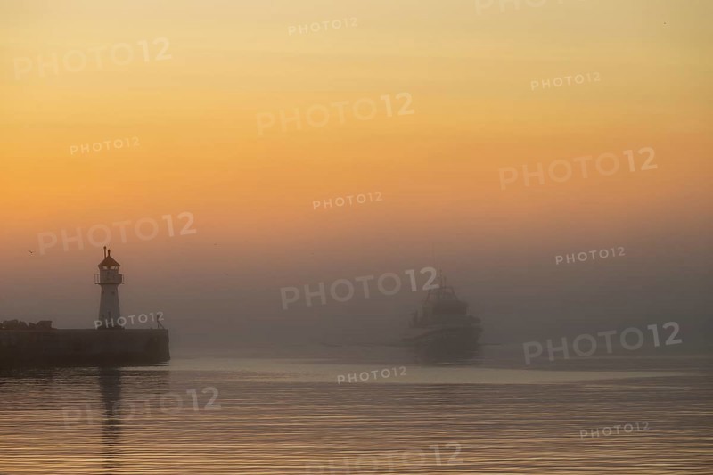 A lighthouse at the entrance to Vardö harbour at sunset with a ship in fog on calm water, Vardø, Finnmark, Norway