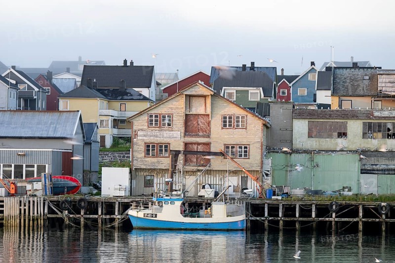 A fishing boat is lying in front of old wooden houses in a quiet harbor, surrounded by morning haze, Vardø, Finnmark, Norway