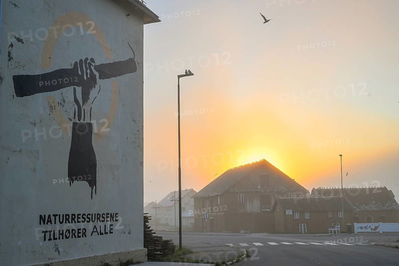 Wall painting with stretched arm and gaff. The inscription says Natural resources belong to everyone. Fishing protet on the Barents Sea against fishing quotas. In the background, the Pomor Museum against the midnight sun, Vardø, Finnmark, Norway