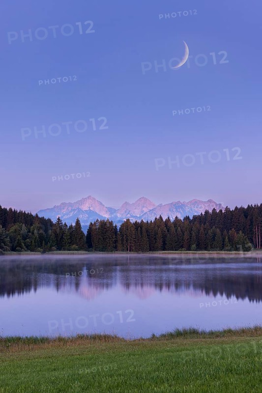 Quiet lake in front of mountains and forest with a narrow moon in the sky, Hegratsrieder See near Füssen, Allgäu Alps, Allgäu, Bavaria, Germany