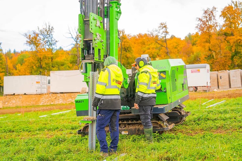 Two workers work with a green machine in front of autumn trees in a meadow, Energiewende, construction of PV open space, Baden-Württemberg, Germany