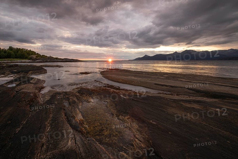 The Vestfjord in Nordland, Norway, offers a dramatic atmosphere near Bodø. Dark clouds are illuminated by the setting sun in shades of red and yellow. The rugged cliffs and mountains are reflected in the quiet fjord, which offers a breathtaking view of Lofoten