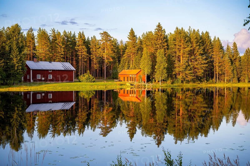 Two red houses are reflected in a quiet lake surrounded by forest under the warm light of the setting sun, Östana, Värmland, Sweden