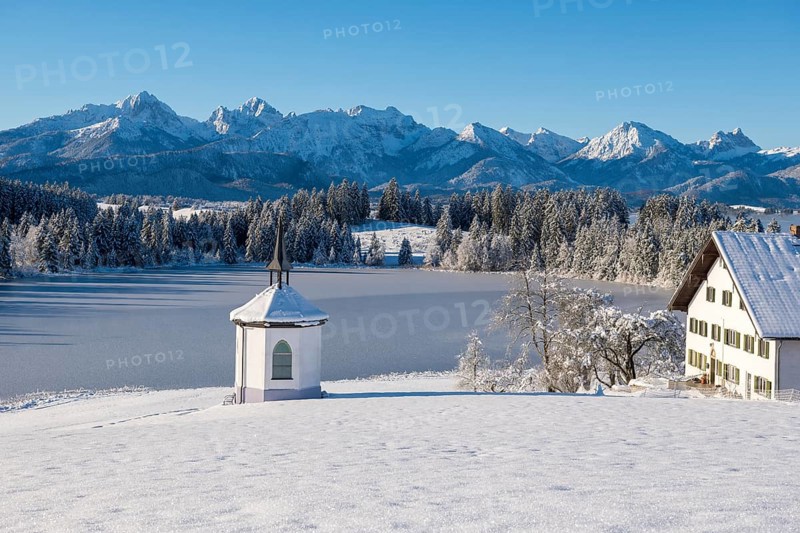 Snowy landscape with chapel, farm and mountains in the background under clear sky, Hegratsrieder See, near Füssen, Ostallgäu, Allgäu, Bavaria, Germany