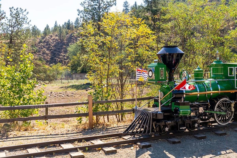 Green miniature train with canadian and british columbian flags stands on tracks in kamloops, british columbia, near a wooden fence and trees, celebrating local and national pride