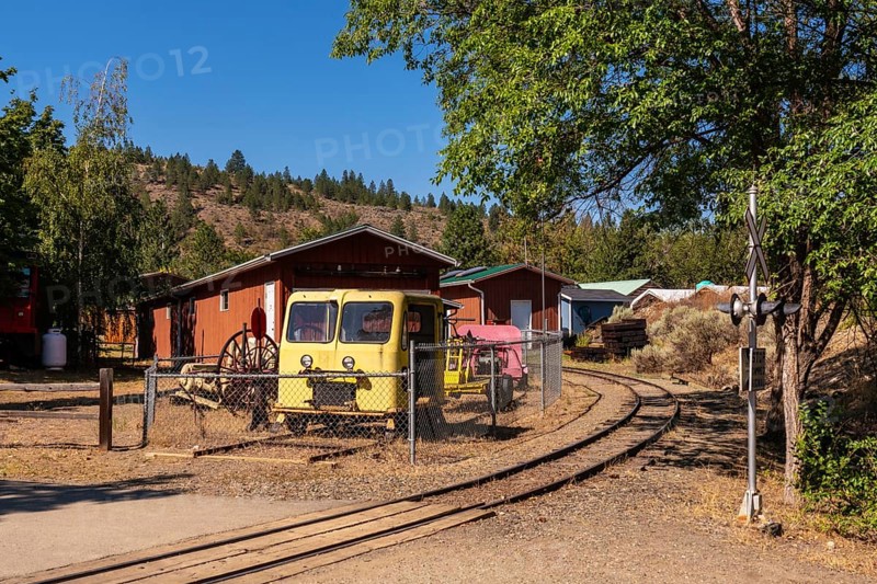 Old yellow locomotive and railway maintenance vehicles resting on a curved rail track in a kamloops yard, surrounded by wooden sheds and forested hills on a sunny day