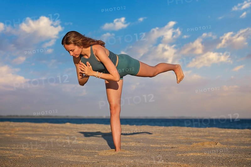 A woman performs Virabhadrasana Warrior 3 pose on the beach during sunset. Her strong stance and focus highlight the beauty of yoga against a tranquil seaside backdrop