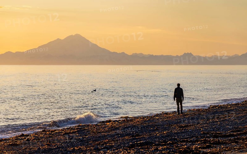 Young man walking along the beach at sunset, view across Cook Inlet to white mountain peaks of Mount Redoubt, Aleutian Range Mountains, Anchor Point, Anchor River State Recreation Area, Alaska, USA