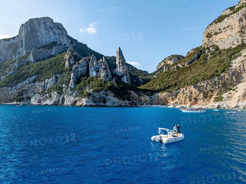 Couple on motorboat off picturesque rocky coast, cliffs with L'Aguglia pinnacle, blue sea and Cala Goloritzé beach, aerial view, Golfo di Orosei, Baunei, Sardinia, Italy