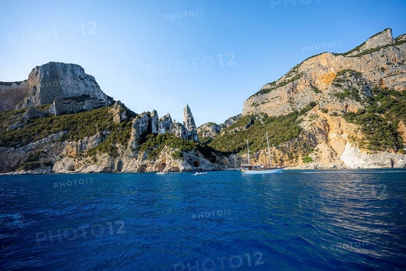 Excursion boat off picturesque rocky coast, cliffs with caves in morning light, L'Aguglia rock pin, blue sea and Cala Goloritzé beach, Golfo di Orosei, Baunei, Sardinia, Italy