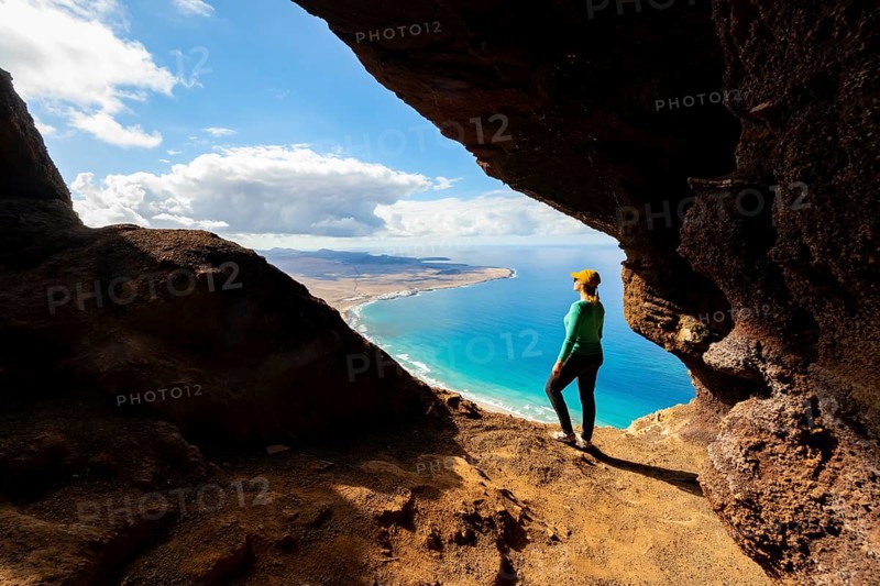Tourist in the Cueva de las Cabras cave, young woman enjoying the view from the Risco de Famara cliff to Famara beach, Playa de Famara with La Calaeta, Lanzarote, Canary Islands, Spain