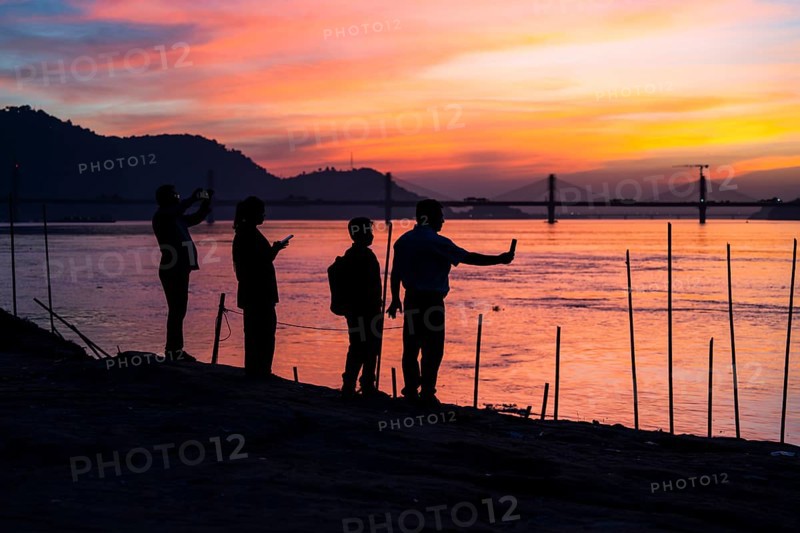 People enjoy the vibrant sunset by the Brahmaputra riverside, capturing photos and moments against the scenic backdrop of a bridge and glowing sky, on November 4, 2025 in Guwahati, India