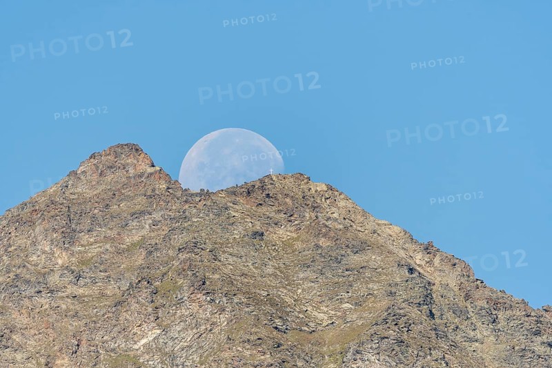 Majestic mountain peak stands tall against vibrant blue sky as large moon rises just behind it. The scene captures the quiet beauty of nature in the early evening light. Grachen, Viege, Valais, Switzerland