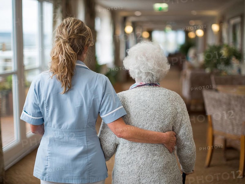A carer assists an elderly woman walking along a bright corridor in a retirement home, nursing home, AI generated