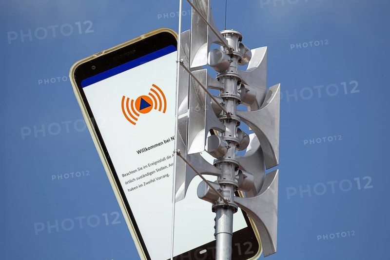 (Photomontage) Close-up of a siren, next to it the open warning app NINA on a smartphone