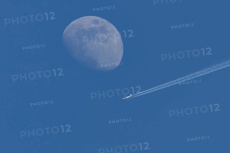 Boeing 737 jet passenger aircraft of Norweign Air with a vapour trial or contrail behind flying in a blue sky passing by the moon, England, United Kingdom
