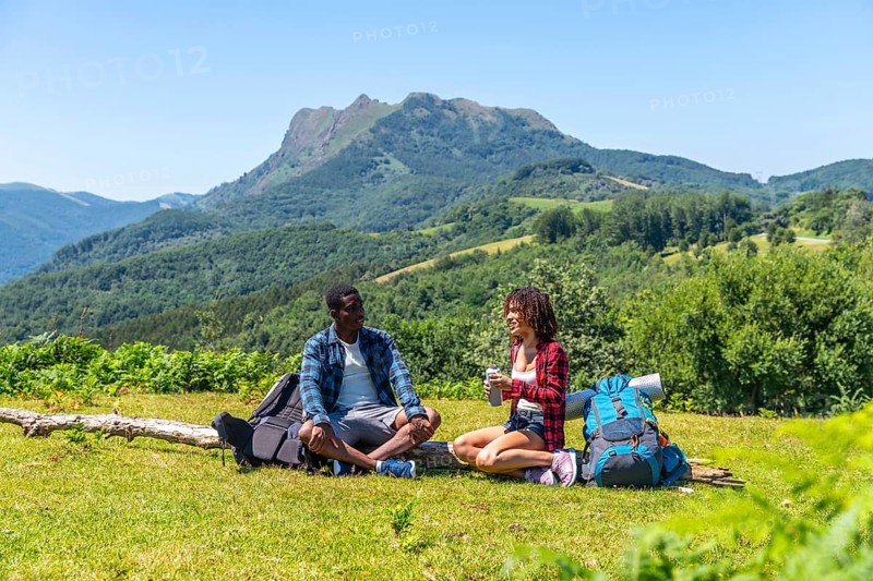 Two hikers are taking a break, sitting on grassy mountaintop and enjoying refreshing drinks with backpacks beside them
