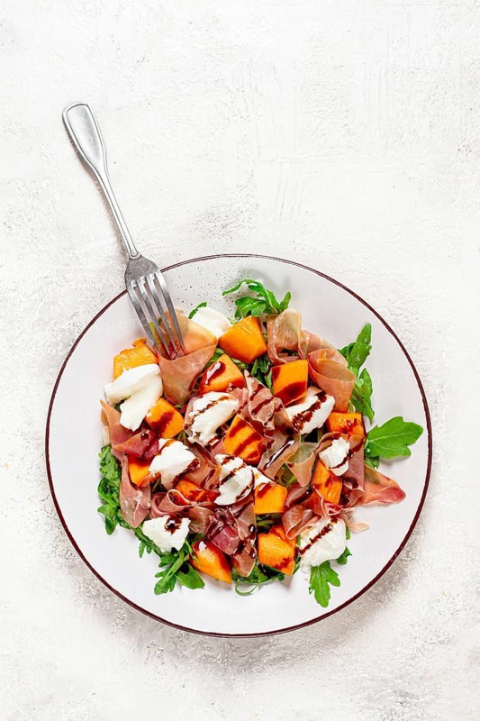 Salad with slices of ripe melon, prosciutto, mozzarella and fresh arugula, decorated with balsamic glaze