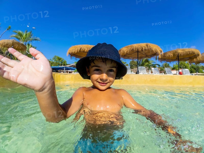 Smiling child in a sun hat waves while playing in a clear swimming pool, surrounded by straw umbrellas and tropical palm trees