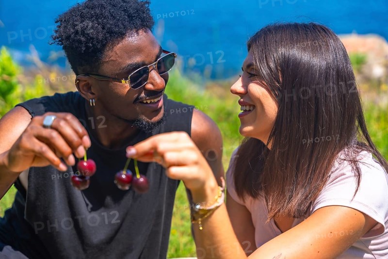 Young man and woman laughing together while sharing fresh cherries, savoring the joy of a sunny summer day in a beautiful outdoor setting