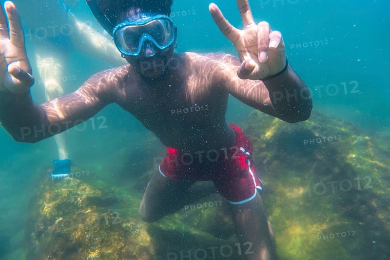 Young man snorkeling underwater, flashing the peace sign with both hands, enjoying the vibrant blue sea and colorful coral reef