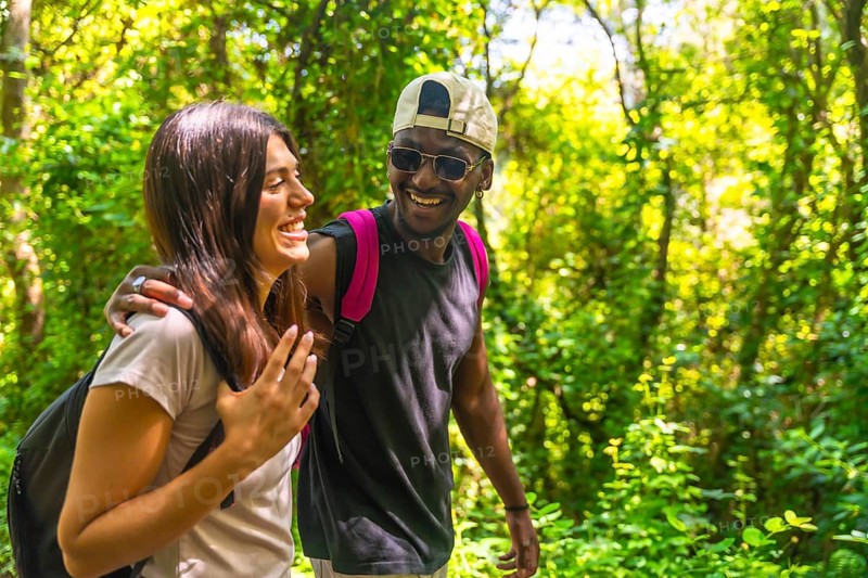 Happy multi ethnic couple enjoying trekking in a green forest, wearing backpacks and casual clothes