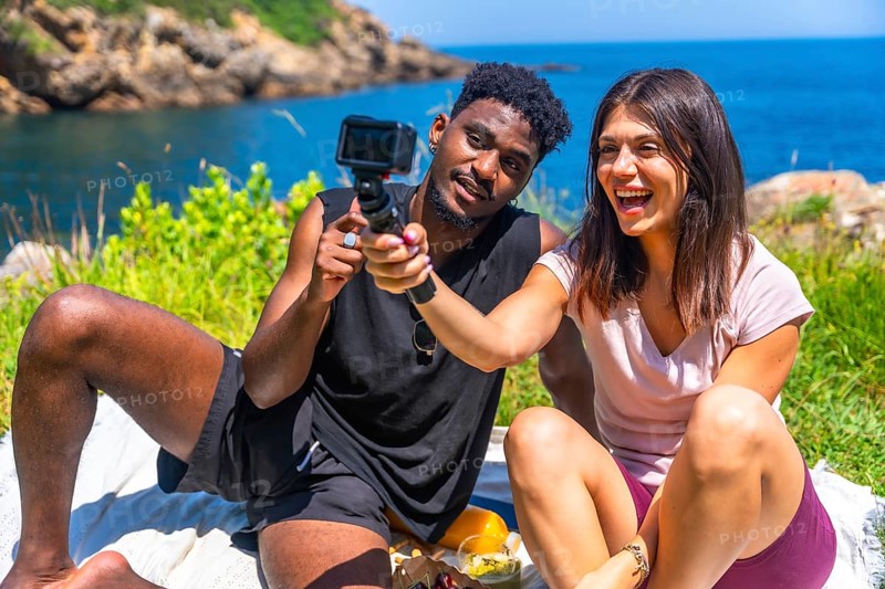 Happy young multi ethnic couple filming with action camera while having picnic by the sea during summer vacation