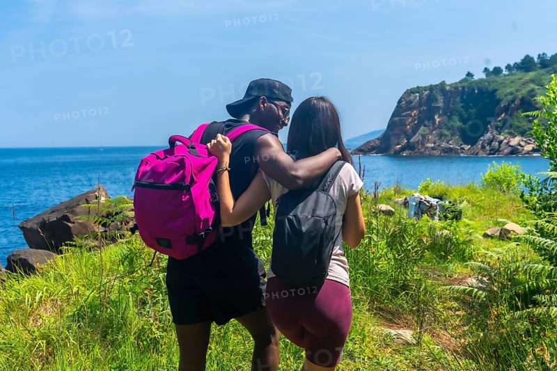 Interracial couple with backpacks walking on a path by the sea, embracing and enjoying the scenic view during a sunny summer day
