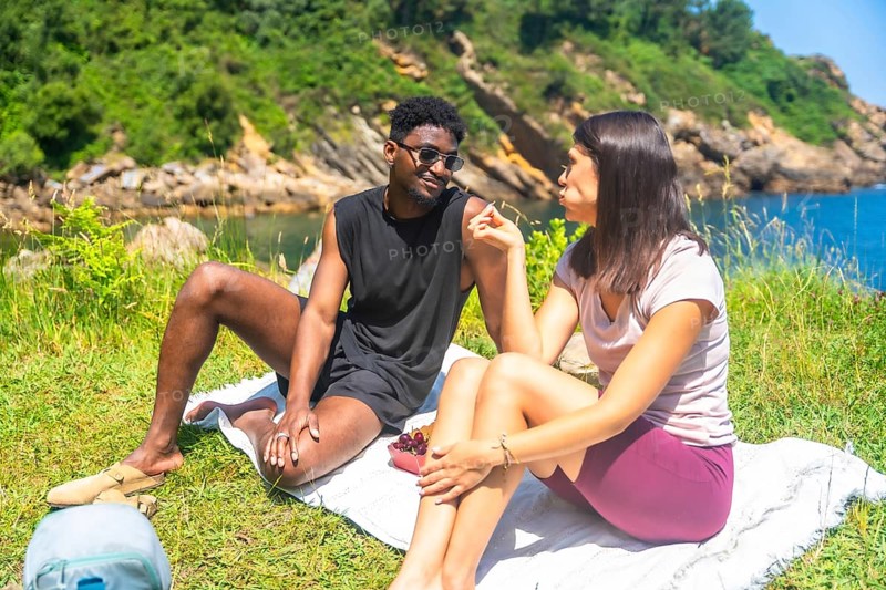 Happy interracial couple sharing grapes during a romantic picnic by the sea on a sunny day, enjoying the beautiful natural scenery