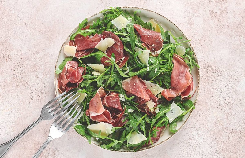 Fresh arugula salad with prosciutto and grated parmesan cheese, on a plate, homemade, no people