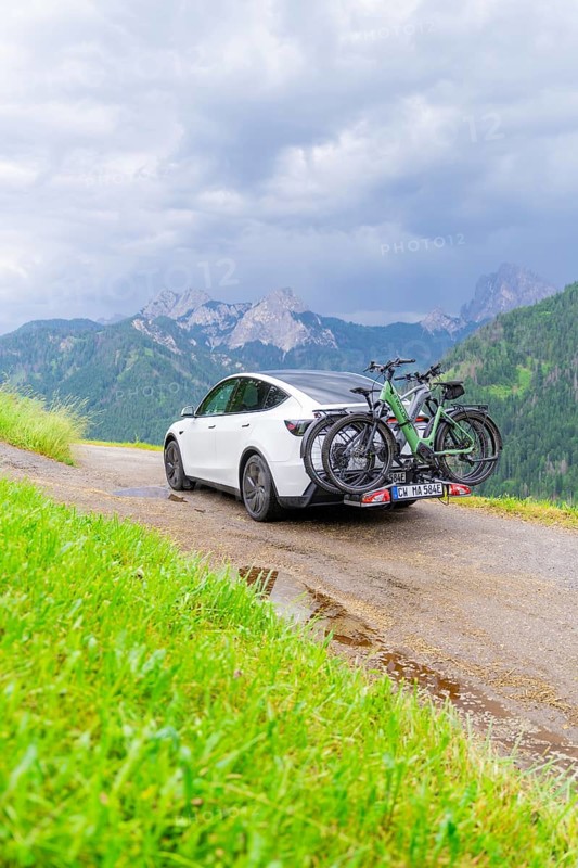 White car with bicycles on mountainous road, surrounded by green landscape and cloudy sky, electric car on holiday, Dolomites, South Tyrol, Dolomites, Italy