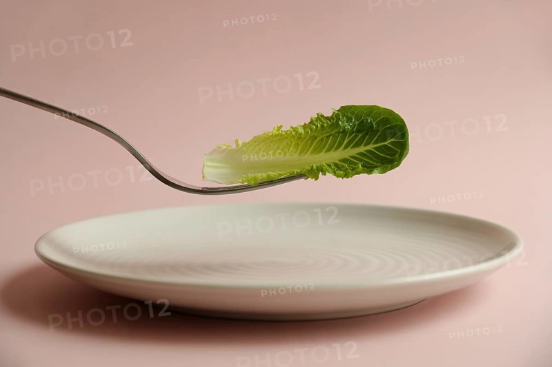 Minimalist close-up of fork holding lettuce leaf above white plate on pink background. Concept of strict diet and unhealthy eating, AI generated