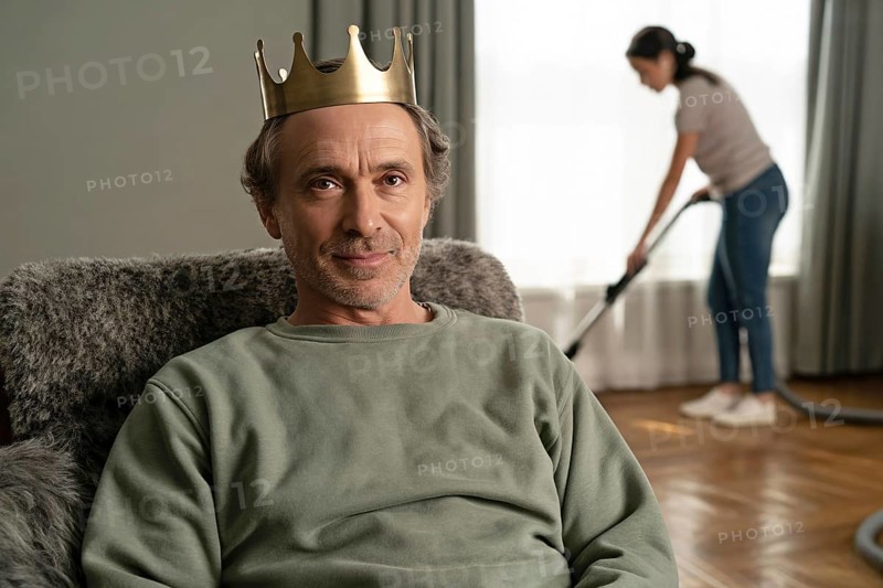 Portrait of middle-aged man with golden crown relaxing on couch while woman vacuums in background. Concept of patriarchal carework imbalance and male privilege. Generative ai, AI generated