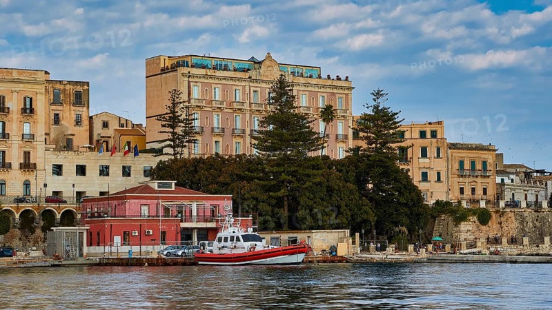 Boats in front of historic buildings and trees on a ponderous coast under a cloudy sky, boat trip around the island of Ortigia, Isola di Ortigia, Syracuse, Sicily, Italy