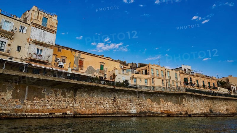 Row of historic houses on a stone seawall under a clear sky, boat trip around the island of Ortigia, Isola di Ortigia, Syracuse, Sicily, Italy