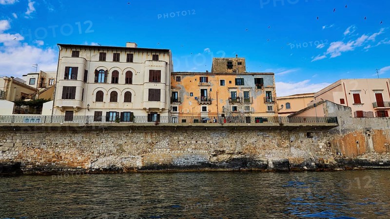Historic buildings along a stone seawall under an expansive blue sky, boat trip around the island of Ortigia, Isola di Ortigia, Syracuse, Sicily, Italy