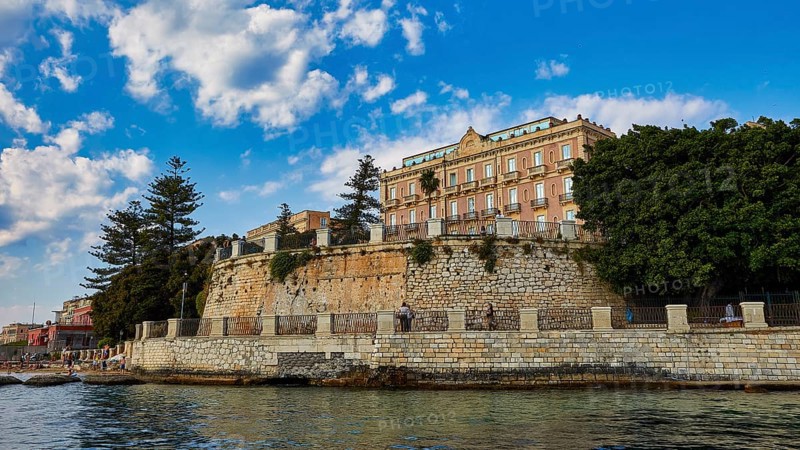 Impressive building on a cliff with lush trees and sunny sky, boat trip around the island of Ortigia, Isola di Ortigia, Syracuse, Sicily, Italy