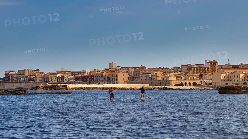 Two paddleboarders on the sea in front of a town with historical buildings under a blue sky, boat trip around the island of Ortigia, Isola di Ortigia, Syracuse, Sicily, Italy
