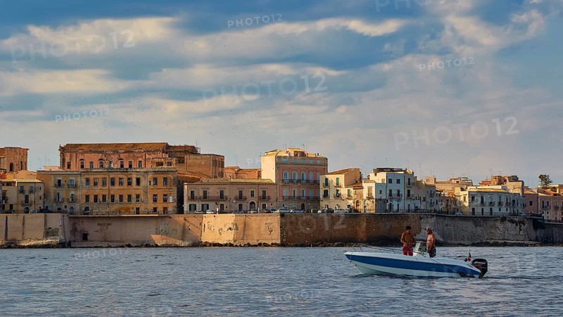 Boat with two people in front of historic coastline and cloudy sky, boat trip around the island of Ortigia, Isola di Ortigia, Syracuse, Sicily, Italy