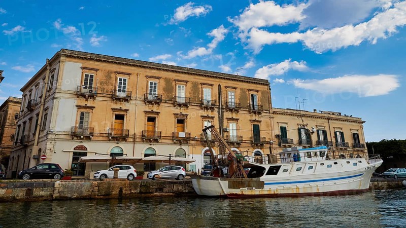 Historic coastal building with parked boat under a blue sky, boat trip around the island of Ortigia, Isola di Ortigia, Syracuse, Sicily, Italy