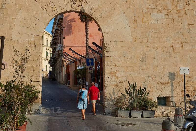People crossing a historic stone city gate surrounded by plants, Ortigia Island, Isola di Ortigia, Historic city centre, Syracuse, Sicily, Italy